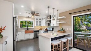 Una cocina moderna con armarios blancos, una isla central con taburetes de bar, un ventilador de techo y vistas al exterior a través de una puerta corrediza.