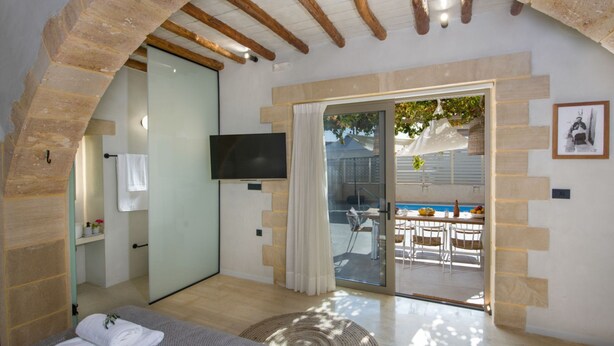 Un dormitorio moderno con un arco de piedra, televisión, balcón con mesa y sillas, y vista a la piscina.