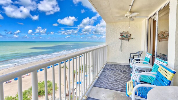 A balcony with a view of the beach and ocean, a ceiling fan, and a row of blue and white chairs. - SandCastle 1804 - Beautiful updates, Breathtaking view, tons of amenities (Indian Shores)