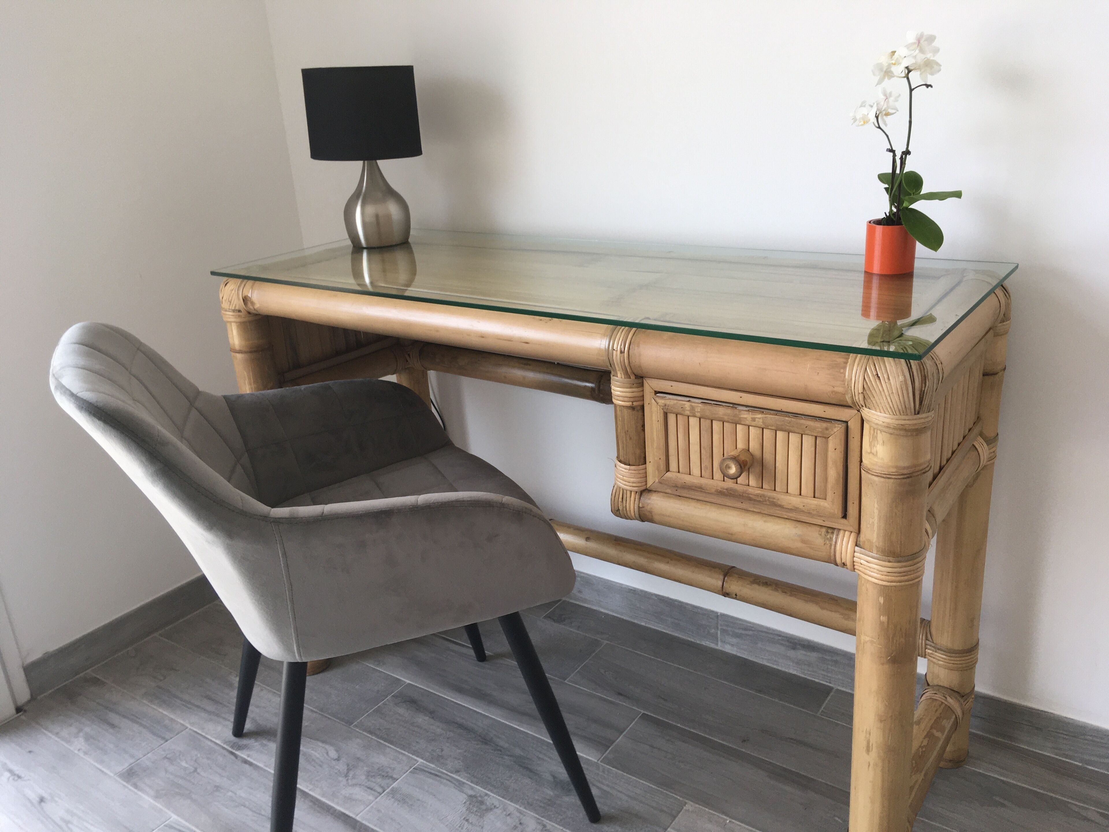Un espace de travail moderne avec un bureau à plateau en verre, une chaise rembourrée de couleur grise, une petite orchidée en pot et une lampe noire.