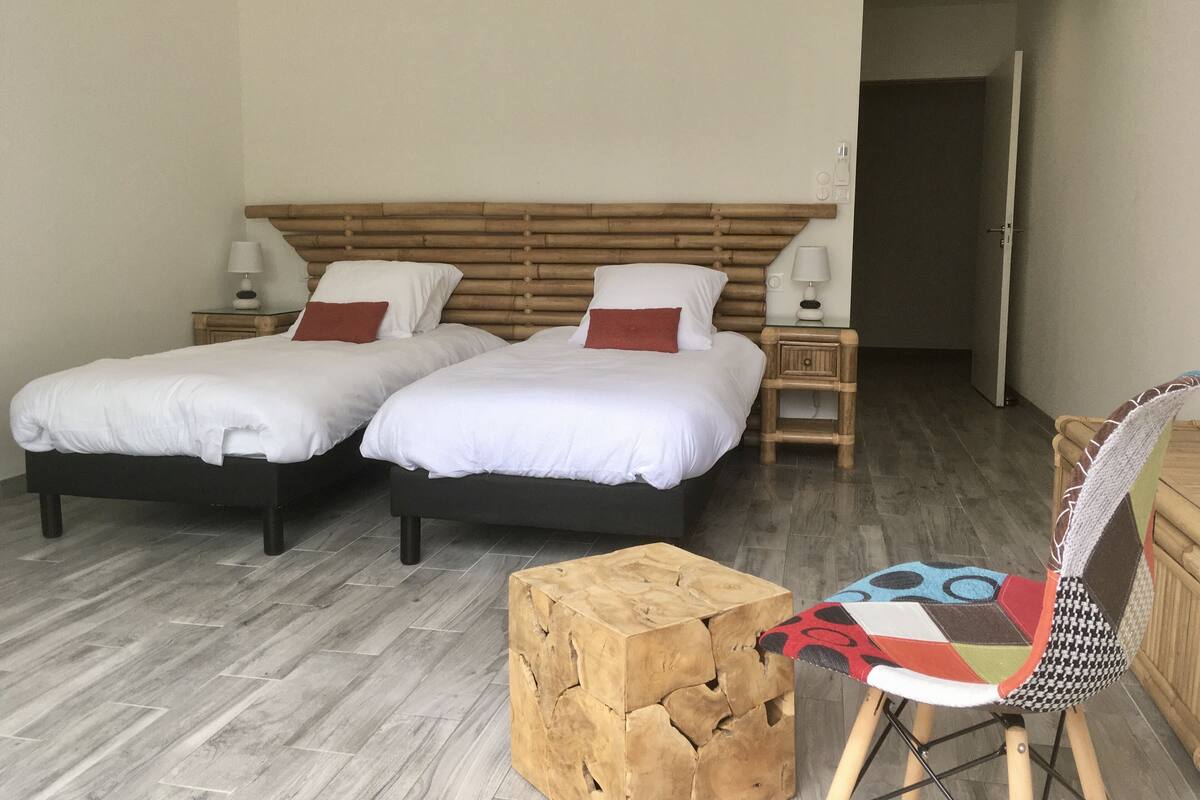 A double room with two beds, wooden headboard, bedside tables with lamps, a wooden stool, and a chair with a colorful cushion.