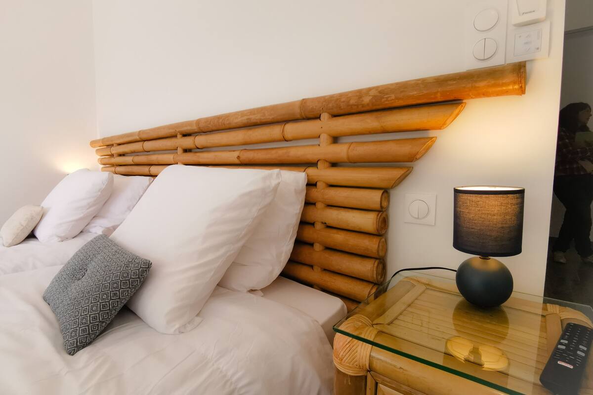 A bed with white bedding and a bamboo headboard, a glass table with a remote control, and a wall-mounted lamp.