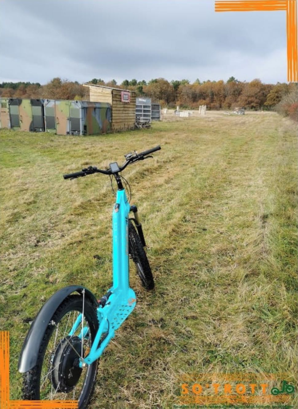 A teal electric bicycle in a grassy field with buildings in the background.