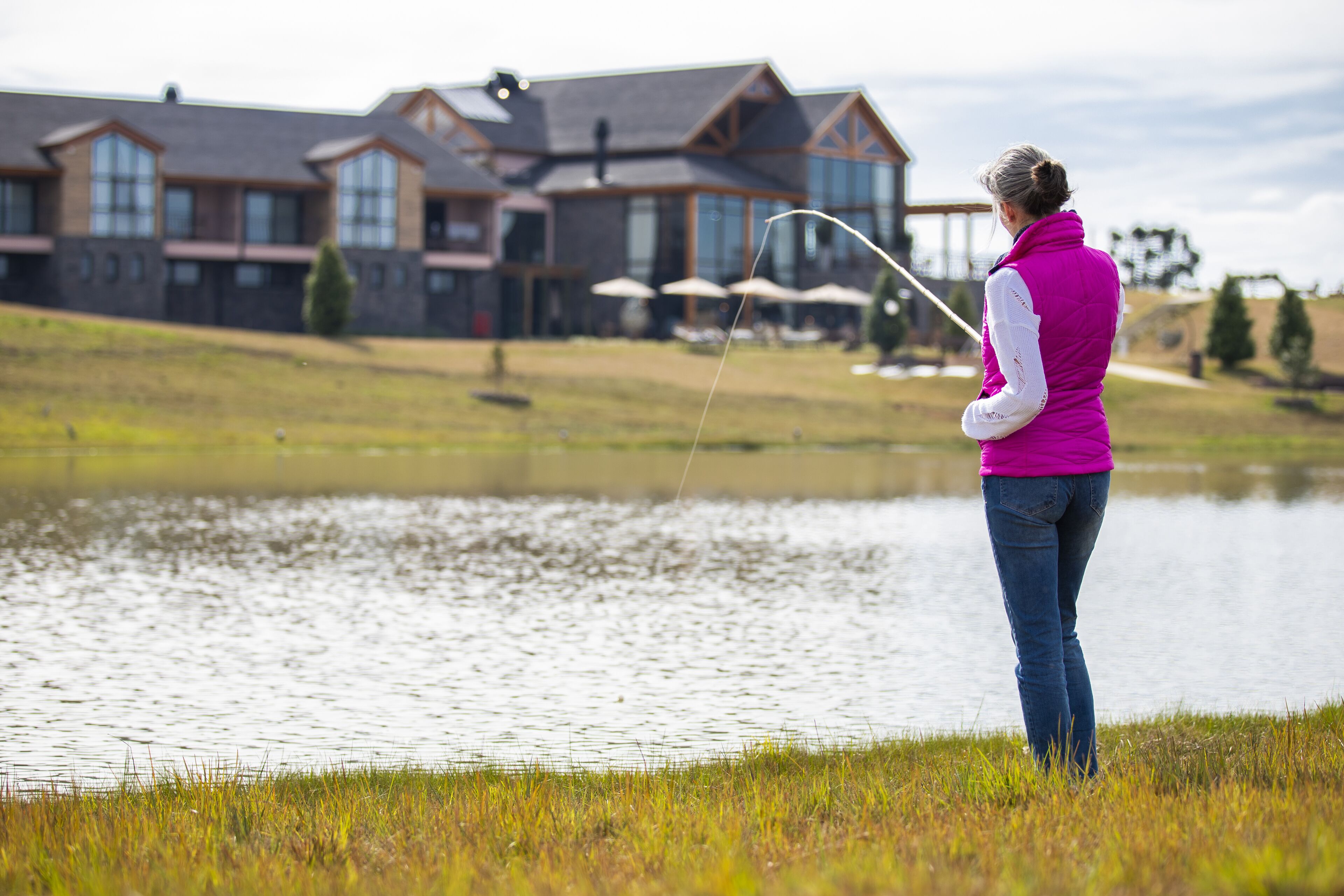 A woman fishing by a calm lake with a house in the background.