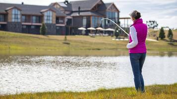 A woman fishing by a calm lake with a house in the background.