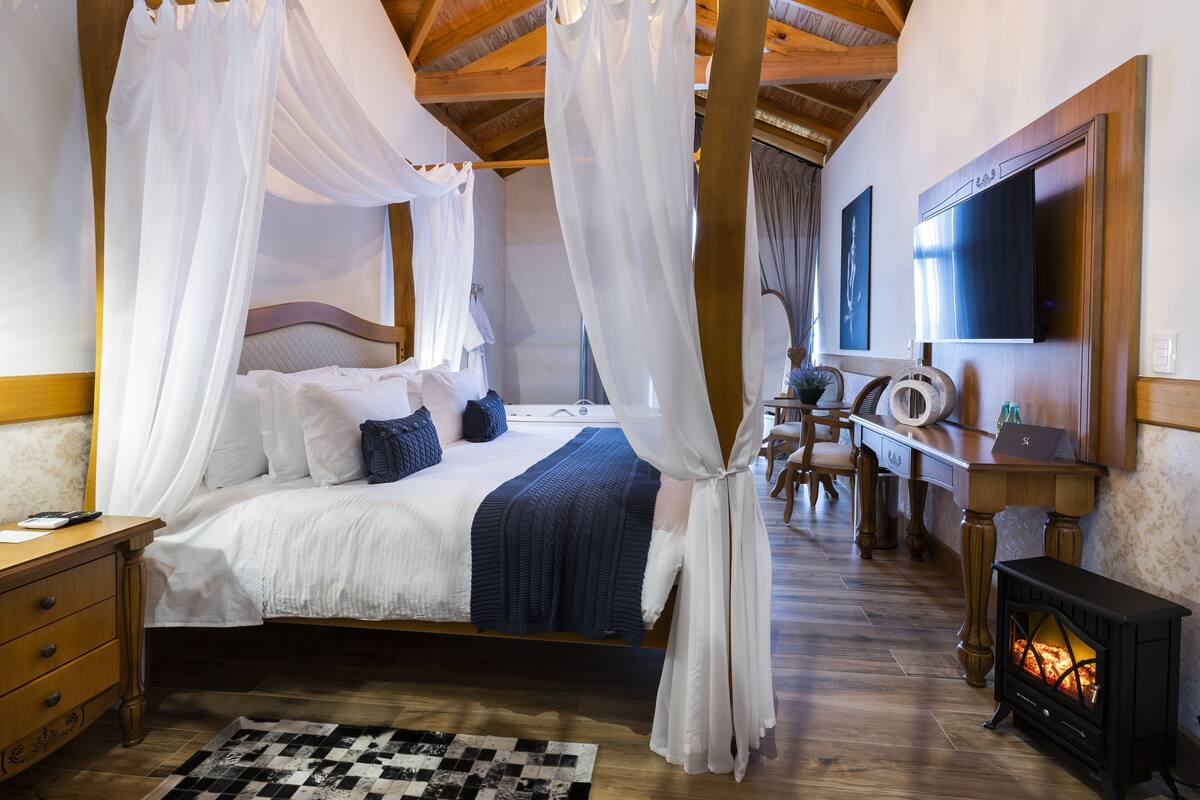 A bedroom with a four-poster bed, a desk with a chair, a television, and a wood-burning stove.
