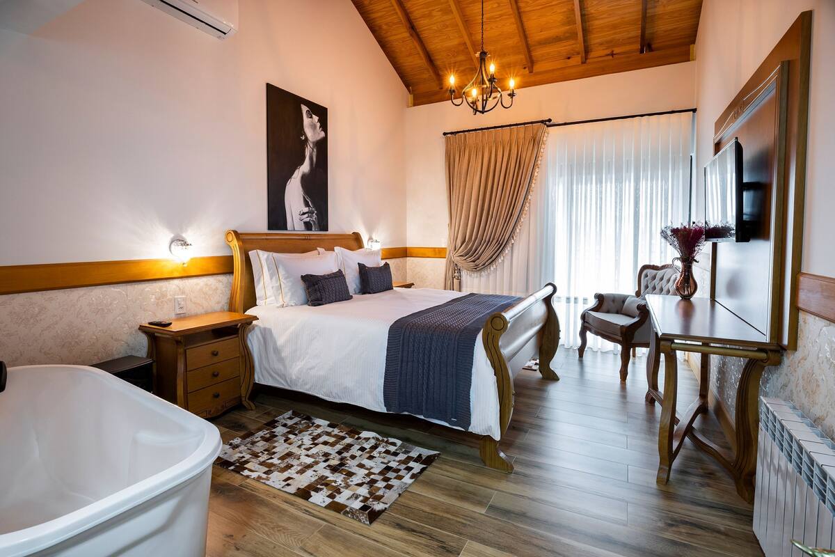 A bedroom with a large bed, a sitting area with a table and chairs, a bathtub, and a mirror.