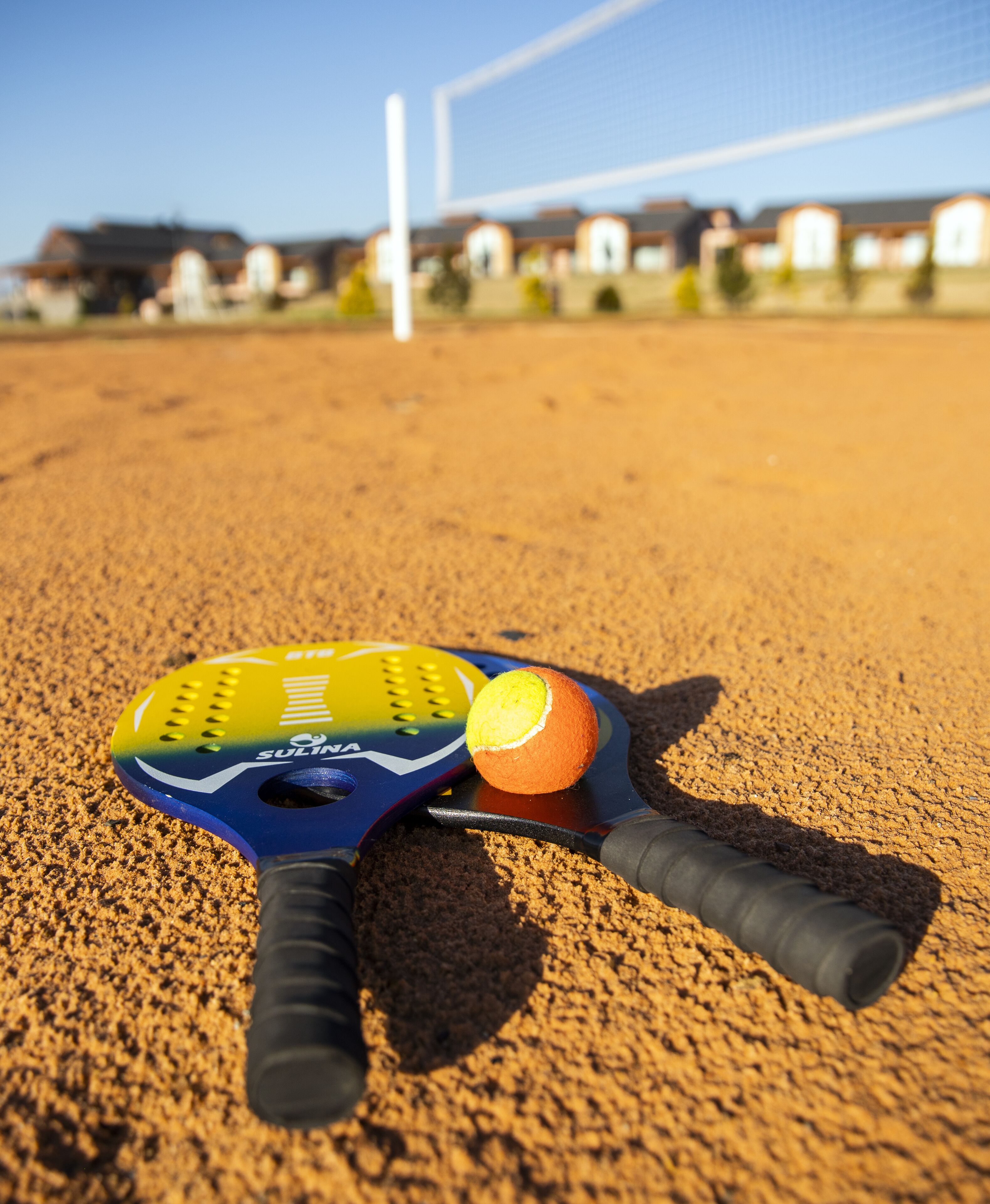 Una raqueta de bádminton y volante sobre arena, con una red y edificios al fondo.