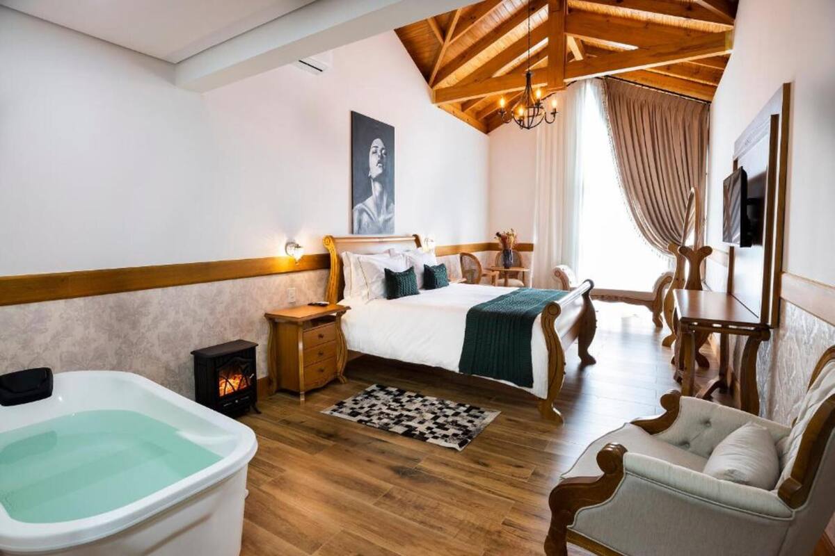 A bedroom with a wooden ceiling, a large bed, a wooden nightstand, a white bathtub, a chair, and a painting on the wall.