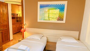 A hotel room with two beds, a wooden wardrobe, a nightstand, a lamp, and a framed artwork on the wall.