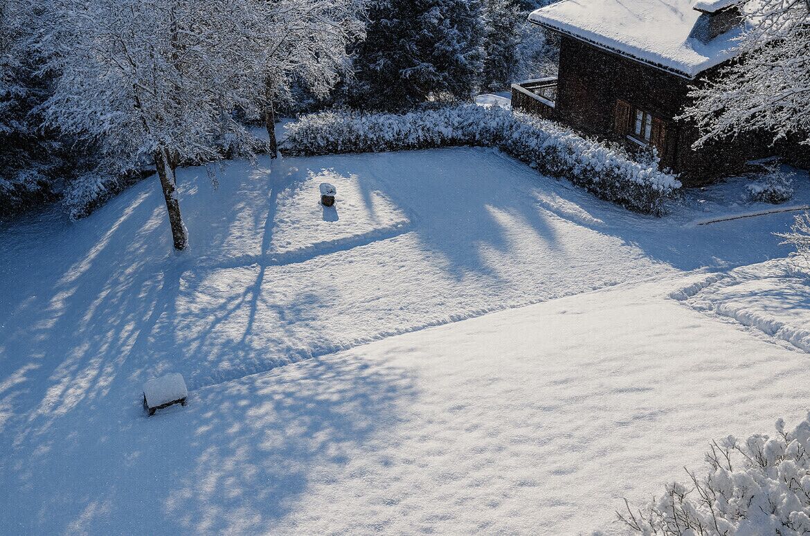 A snow-covered yard with a wooden bench and a house in the background.
