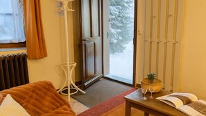 A cozy room with a brown sofa, a wooden table with a book and glasses, a floor lamp, and a view of snow outside.