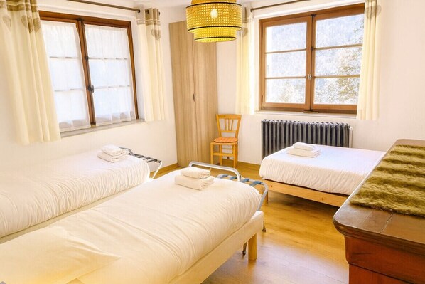 A hotel room with two beds, a wooden chair, a radiator, and a window with curtains.