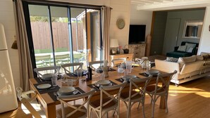 A dining area with a wooden table set for six, surrounded by white chairs with woven seats. A television is mounted on the wall above the sofa in the adjacent living area. - Sumptuous Cap Ferret Style House (La Teste-de-Buch)