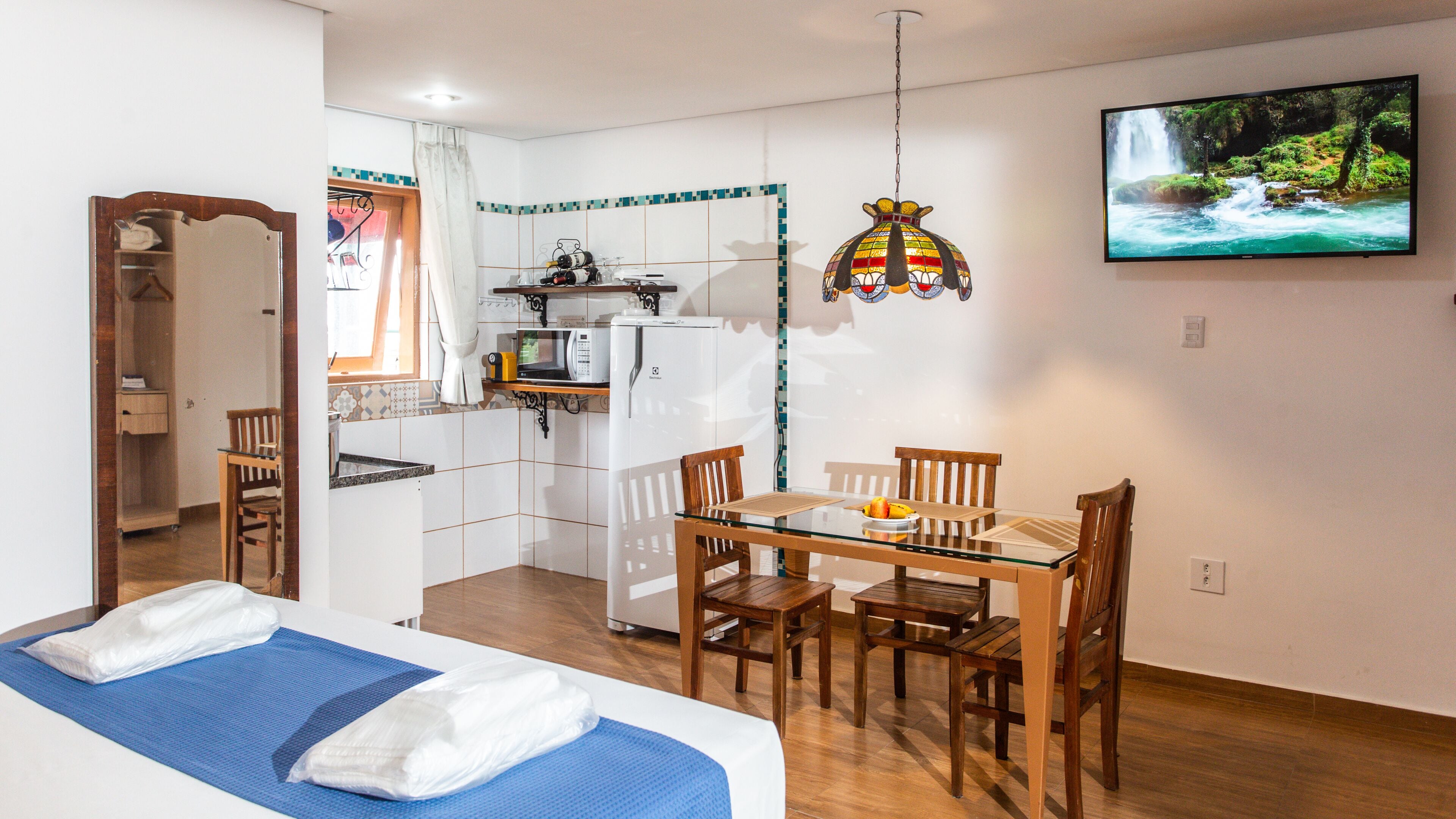 A compact hotel room with a bed, a dining table with chairs, a kitchenette, and a TV on the wall.