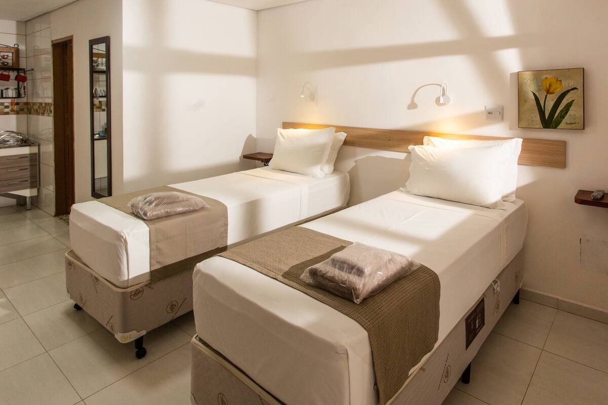 Quarto de hotel com duas camas de solteiro, cabeceira de madeira, mesa de cabeceira com abajur, um quadro na parede e um banheiro visível por uma porta aberta.
