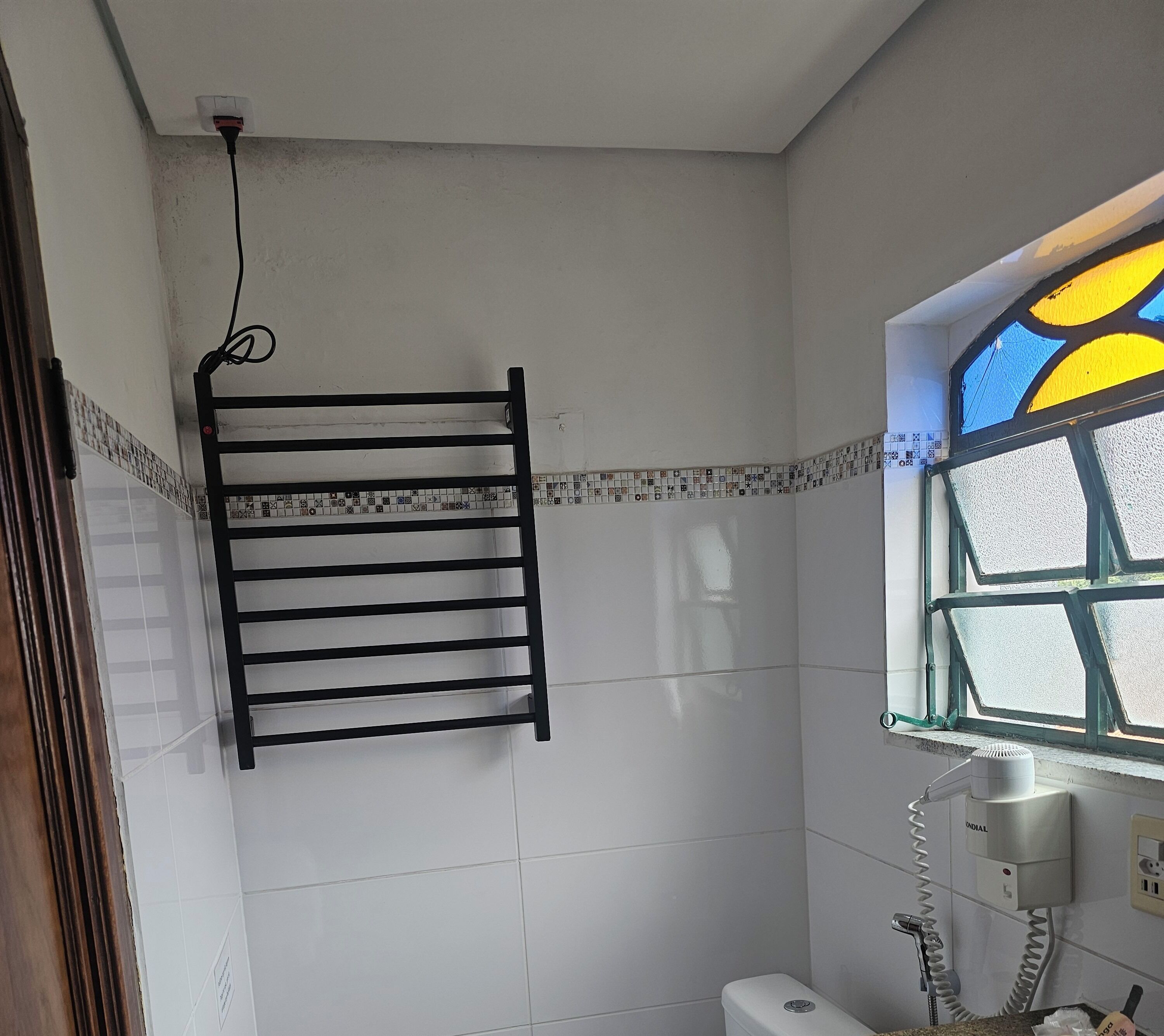A bathroom with white tiled walls, a black towel rack, a window with stained glass, and a toilet.