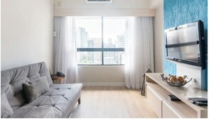 Een moderne woonkamer met een grijze bank, een aan de muur bevestigde televisie en een raam met gordijnen.