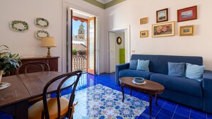 A living room with a blue sofa, a dining table with chairs, a patterned rug, and a view of a building through an open door. - Stefania Flat- Amalfi centre (Amalfi)