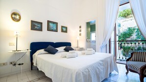 A bedroom with a large bed, a small table, a chair, and a balcony with a view of greenery. - Stefania Flat- Amalfi centre (Amalfi)