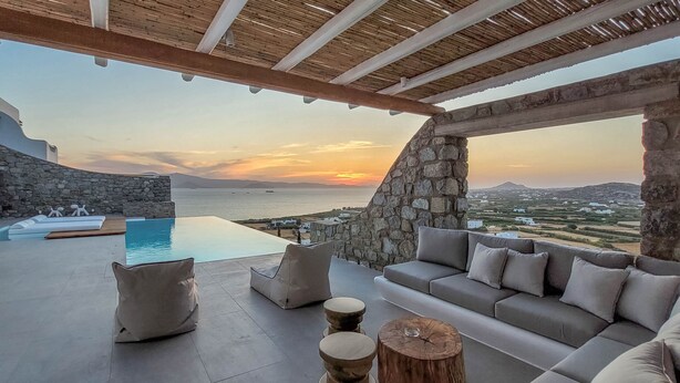 Une terrasse avec un mur en pierre, une piscine et une vue sur la mer au coucher du soleil.