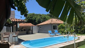 En swimmingpool med solstolar, ett område med grillplats och en liten byggnad med ett tak täckt av kakel.