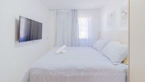 Une chambre à coucher avec un lit, un téléviseur à écran plat, des rideaux blancs et un tableau encadré au mur.