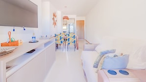 Une pièce moderne et bien éclairée, avec un canapé blanc, une table à manger recouverte d’une nappe à motifs floraux bleus et jaunes, et un téléviseur à écran plat fixé au mur.