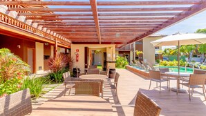Une terrasse extérieure avec des tables rondes et des chaises en osier, une piscine et une allée couverte.