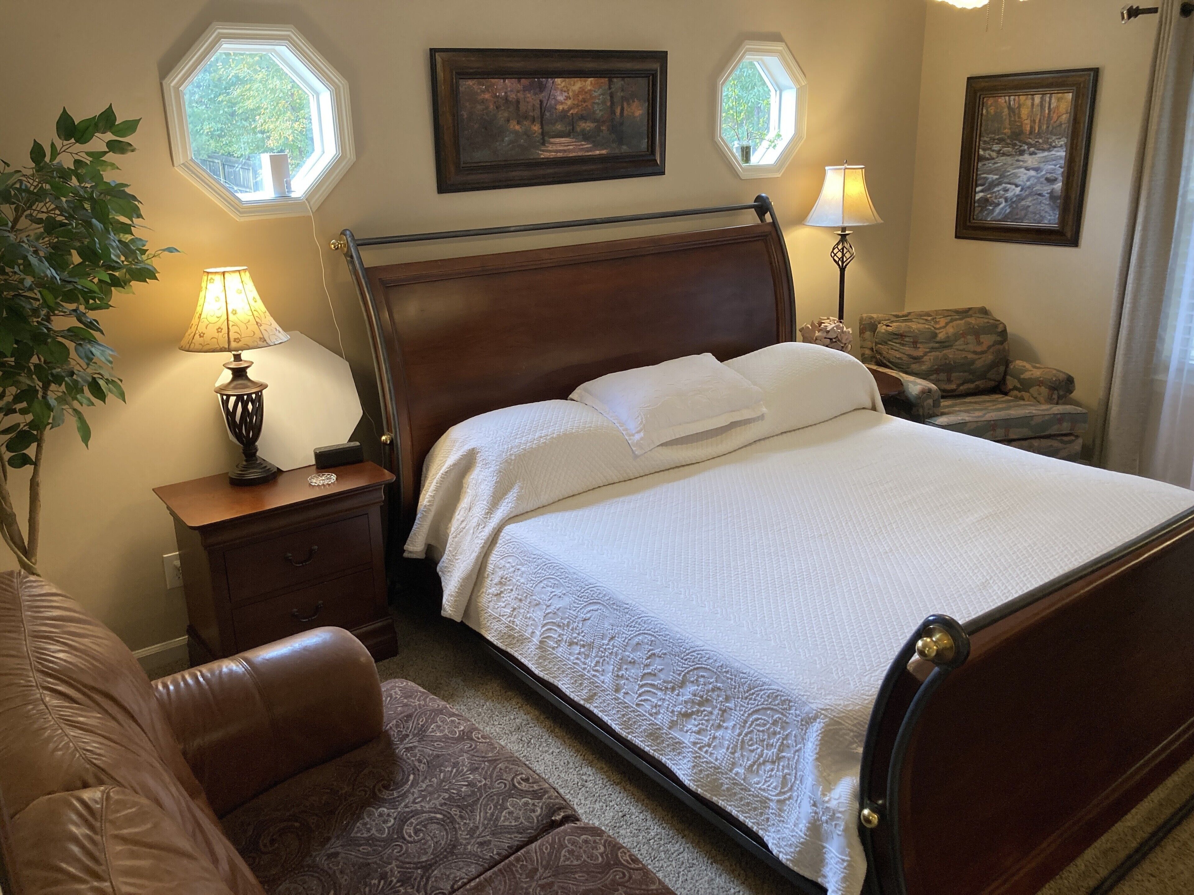 A neatly arranged bedroom with a wooden bed, a nightstand with a lamp, a chair, and two framed pictures on the wall.
