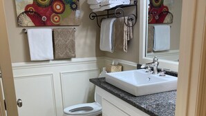 Un baño con inodoro blanco, un lavabo con encimera de granito y un espejo sobre el lavabo. Hay toallas colgadas en un perchero y un cuadro decorativo en la pared.