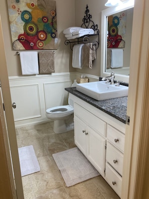 Un baño con inodoro blanco, un lavabo con encimera de granito y un espejo sobre el lavabo. Hay toallas colgadas en un perchero y un cuadro decorativo en la pared.