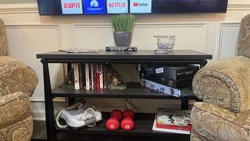 A living room with a flat-screen TV displaying various streaming services, a bookshelf with books and a chess set, and a pair of dumbbells on a shelf.