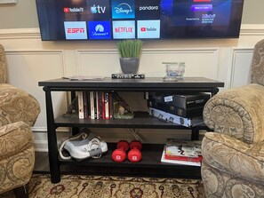 Una sala de estar con una televisión de pantalla plana mostrando diversos servicios de streaming, una estantería con libros y un juego de ajedrez, y un par de mancuernas sobre un estante.
