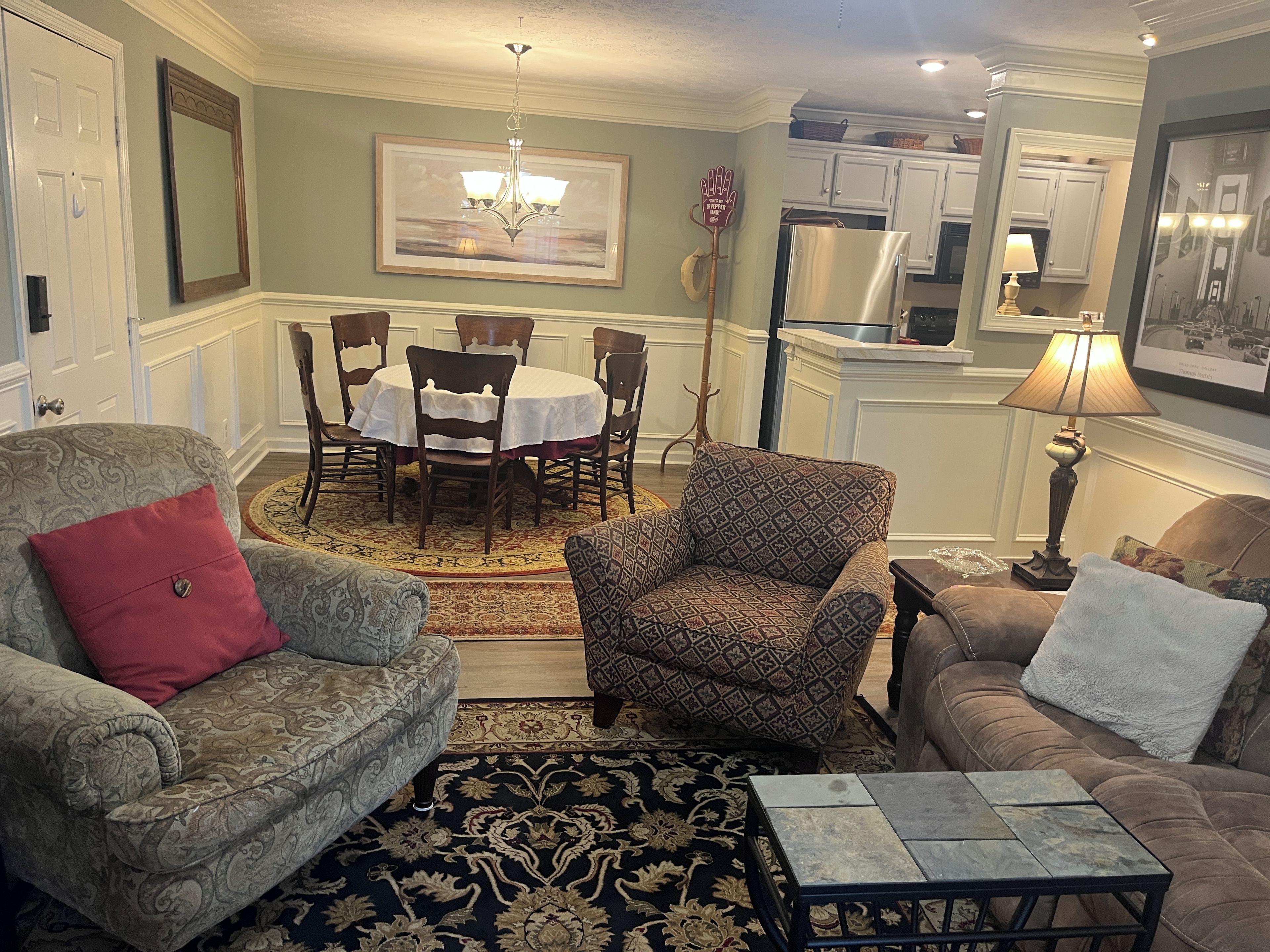 A living room with a patterned armchair, a sofa, a coffee table, a dining table with chairs, and a kitchen area in the background.