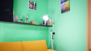 Um quarto com parede verde, sofá amarelo, uma estante com livros e objetos decorativos, e uma luminária de mesa.