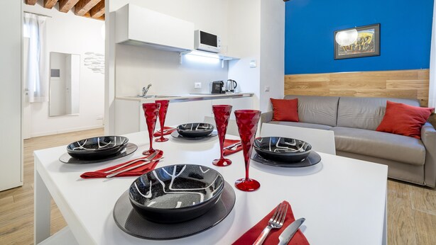 Un comedor moderno con una mesa blanca dispuesta para cuatro personas, copas rojas y un sofá gris con cojines rojos al fondo.