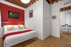 Quarto com cama, parede de destaque vermelha, pintura e área de jantar com mesa e cadeiras.