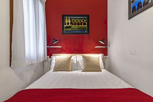 Un letto rifatto con lenzuola bianche e due cuscini fantasia, una parete rossa a contrasto e un'opera d'arte incorniciata sopra il letto.