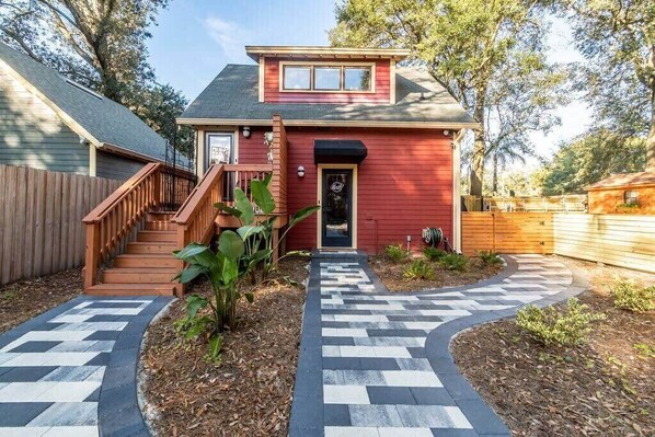 A red house with a wooden staircase leading to the front door, surrounded by a garden with a patterned walkway.
