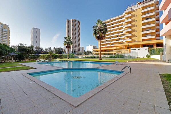 A swimming pool surrounded by a tiled patio area, with a tall building in the background.