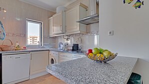 Une cuisine avec des meubles blancs, un plan de travail en granit, un lave-vaisselle, une machine à laver et un panier à fruits.