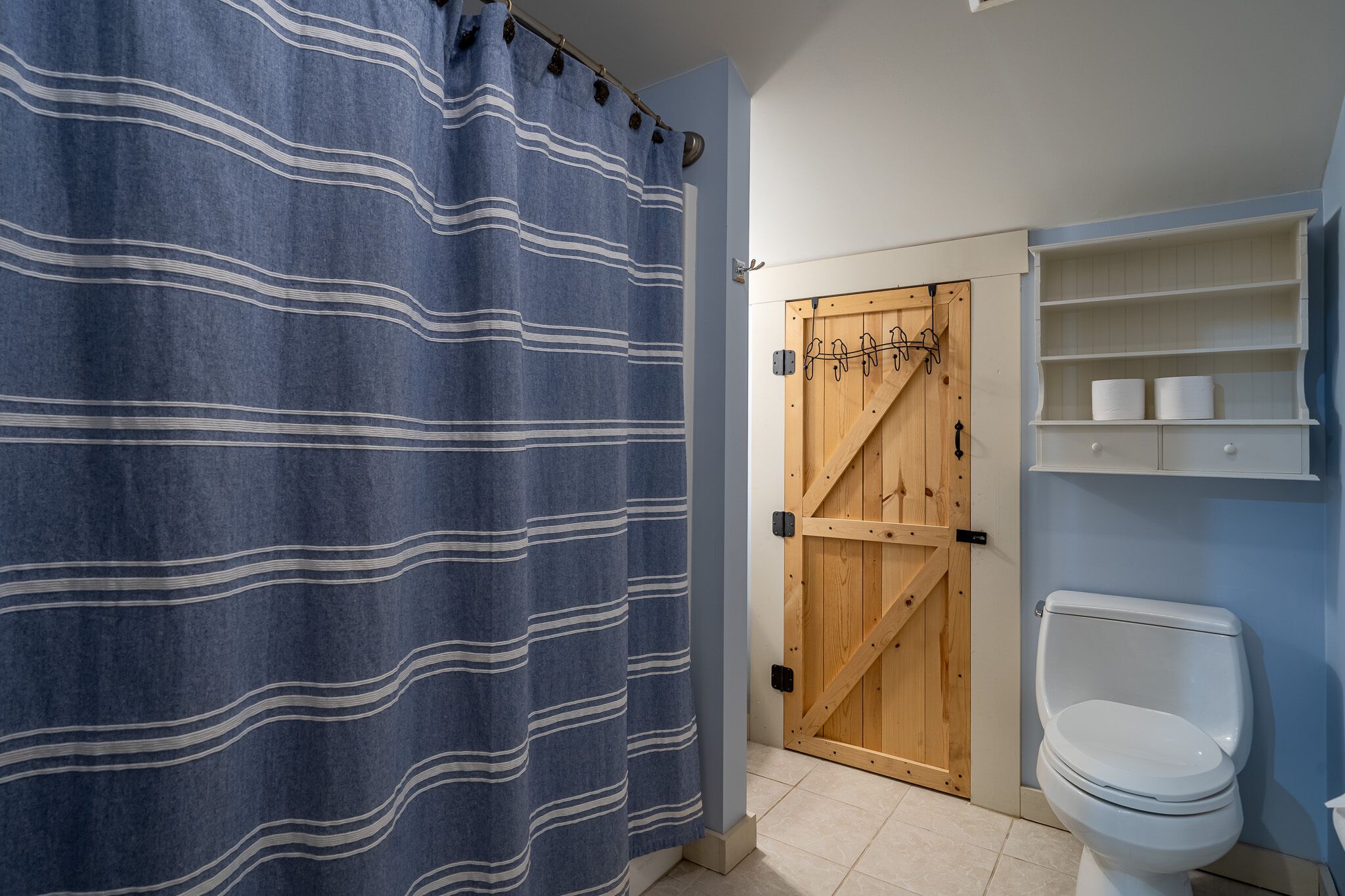 Ein Badezimmer mit blauer, gestreifter Duschvorhang, einer Holztür, einer weißen Toilette und integrierten Regalen.