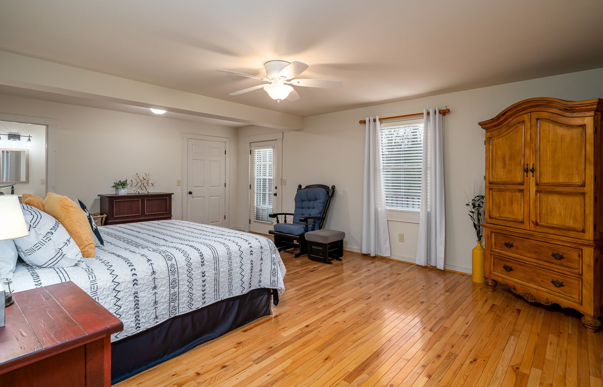 Une chambre avec un plancher en bois, un lit avec une literie à motifs, un fauteuil à bascule, une armoire en bois et un ventilateur de plafond.