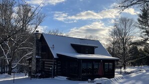 Una cabaña de troncos con techo nevado, rodeada de árboles y suelo cubierto de nieve.