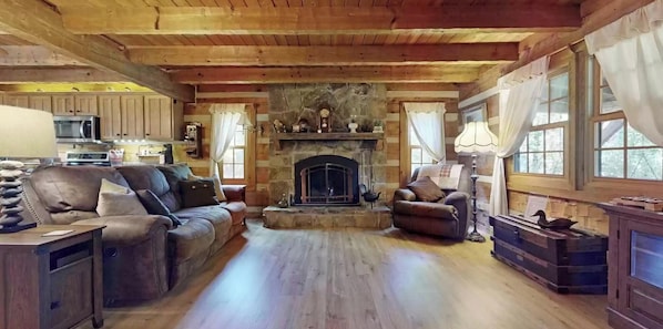 A cozy cabin interior with a stone fireplace, wooden ceiling, and a living area with leather sofas and a dining table.