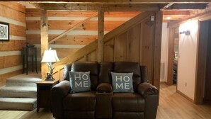 Un acogedor interior de cabaña con un sofá de cuero marrón, dos almohadas con el texto 'HOME', una lámpara y un cuadro en la pared.