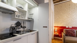 A compact kitchen with white cabinets, a black induction cooktop, and a coffee machine. A small dining area with a red wall and a sofa is visible. - Ca' dell'Arciere - Superior Apartment in the Heart of Venice (Venezia)