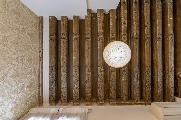 Una habitación con columnas de madera ornamentadas, un papel tapiz estampado y una lámpara redonda de fibras naturales.