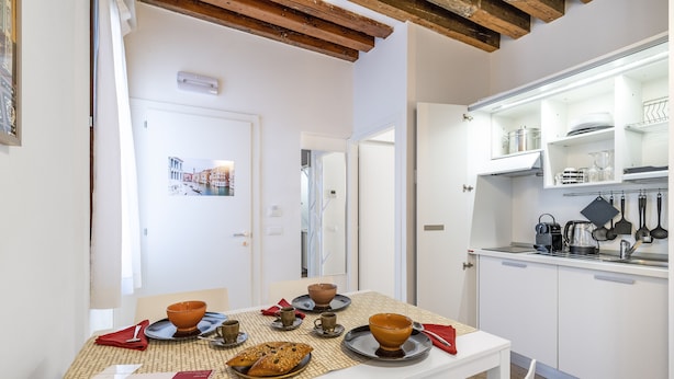 A dining area with a table set for two, a kitchen with white cabinets and stainless steel appliances, and a framed picture on the wall. - Ca' dell'Arciere - Superior Apartment in the Heart of Venice (Venezia)